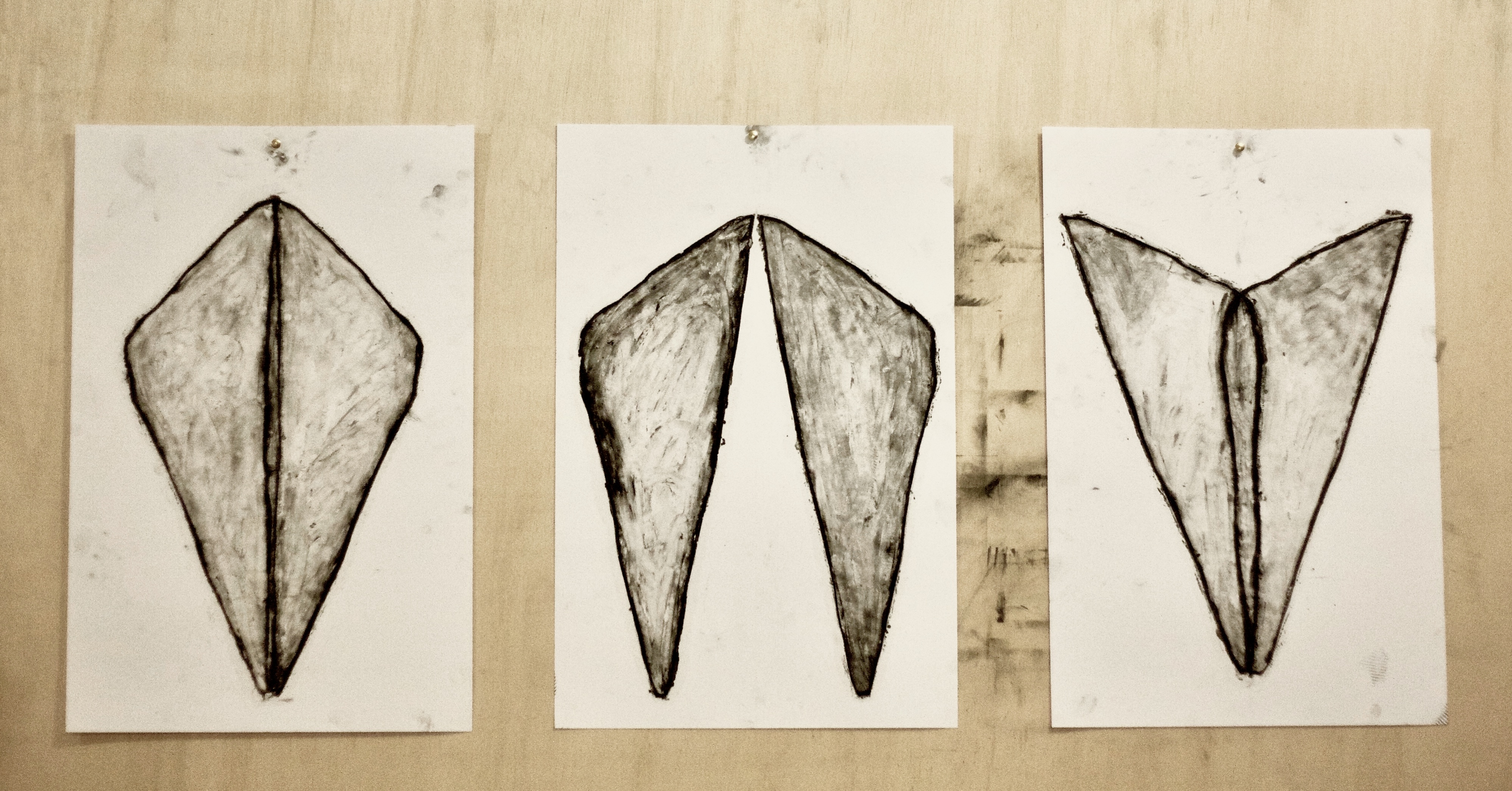 Três desenhos de formas simétricas em papel sobre parede de madeira.